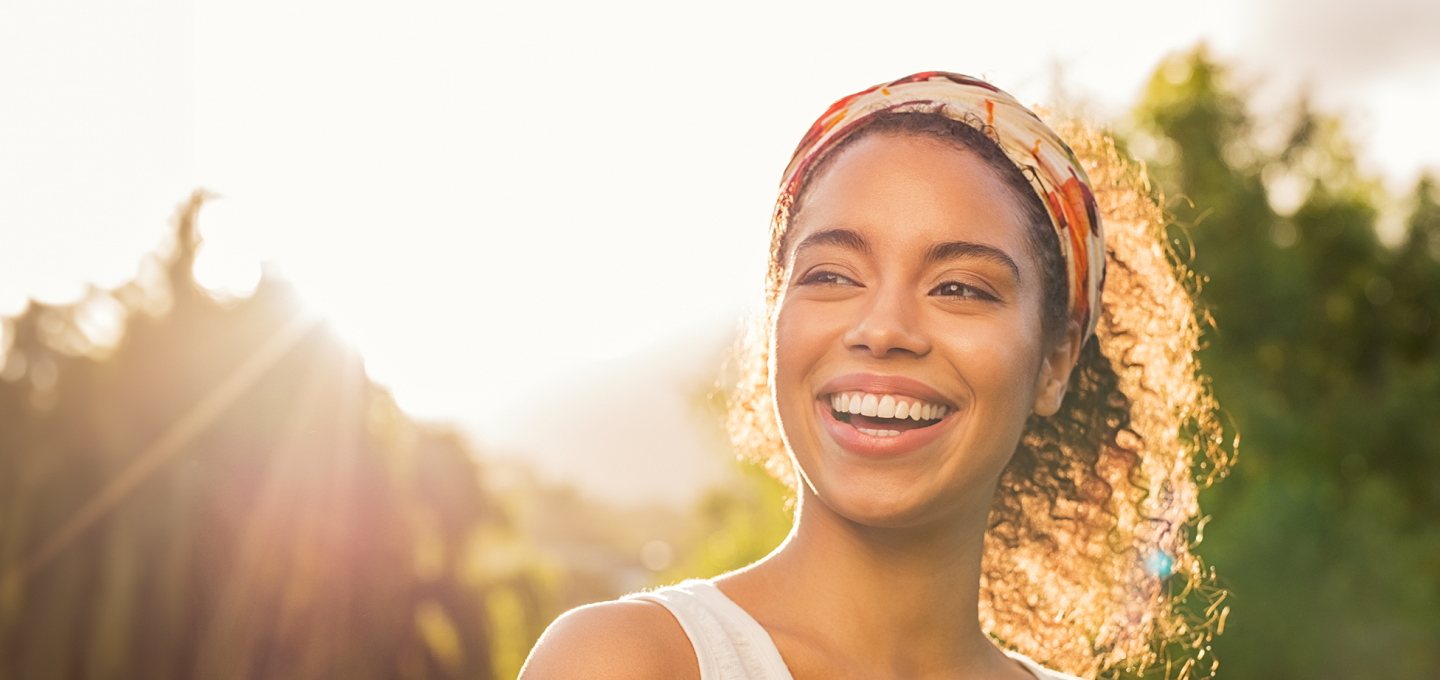 Imagem de uma mulher preta, com cabelos pretos cacheados em um local com árvores. Ela tem um sorriso bonito, está feliz e passa muita autoestima.