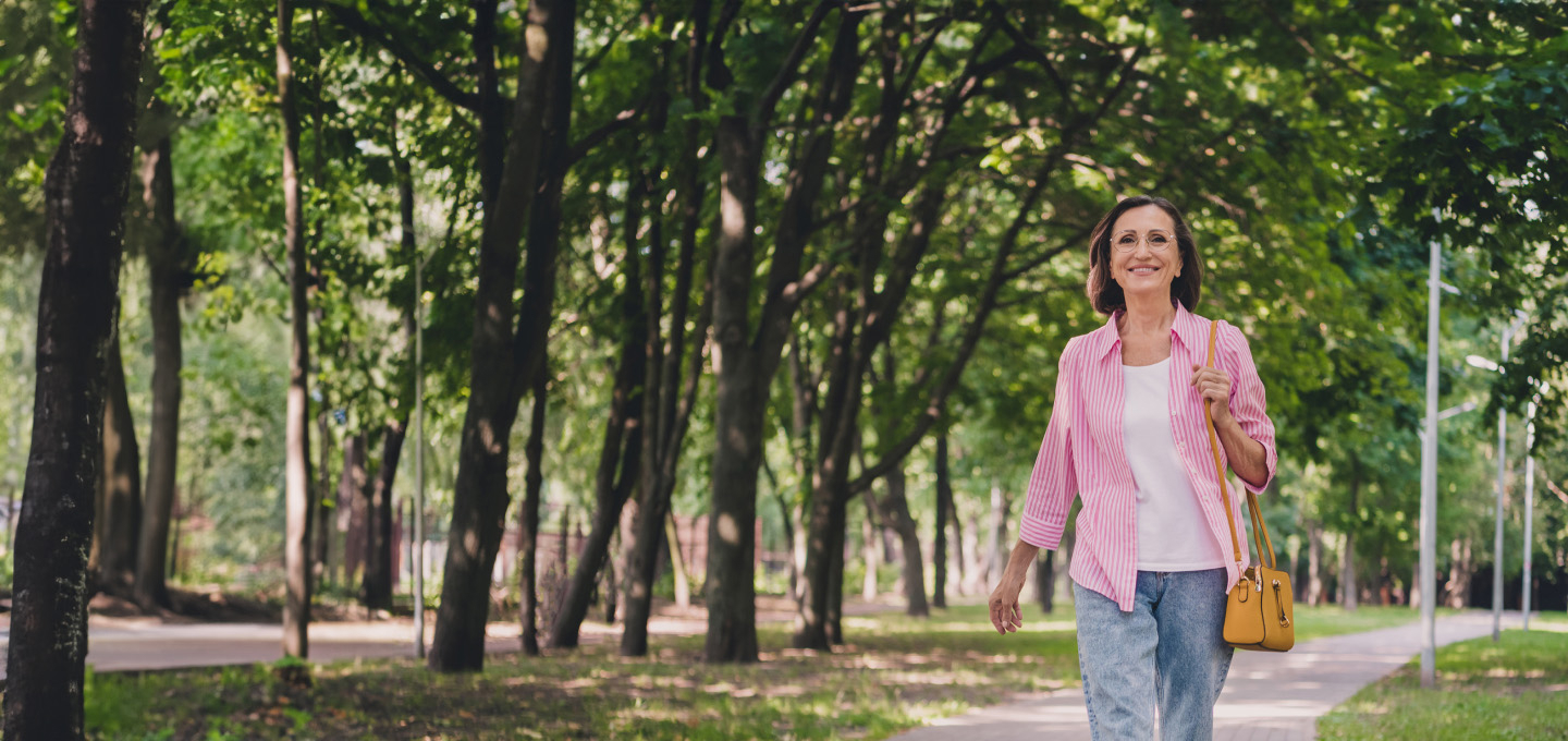 Imagem de uma mulher de meia idade andando em um parque durante o dia. Ela veste camisa rosa, jeans azul e carrega uma bolsa marrom com ar de proteção e tranquilidade.