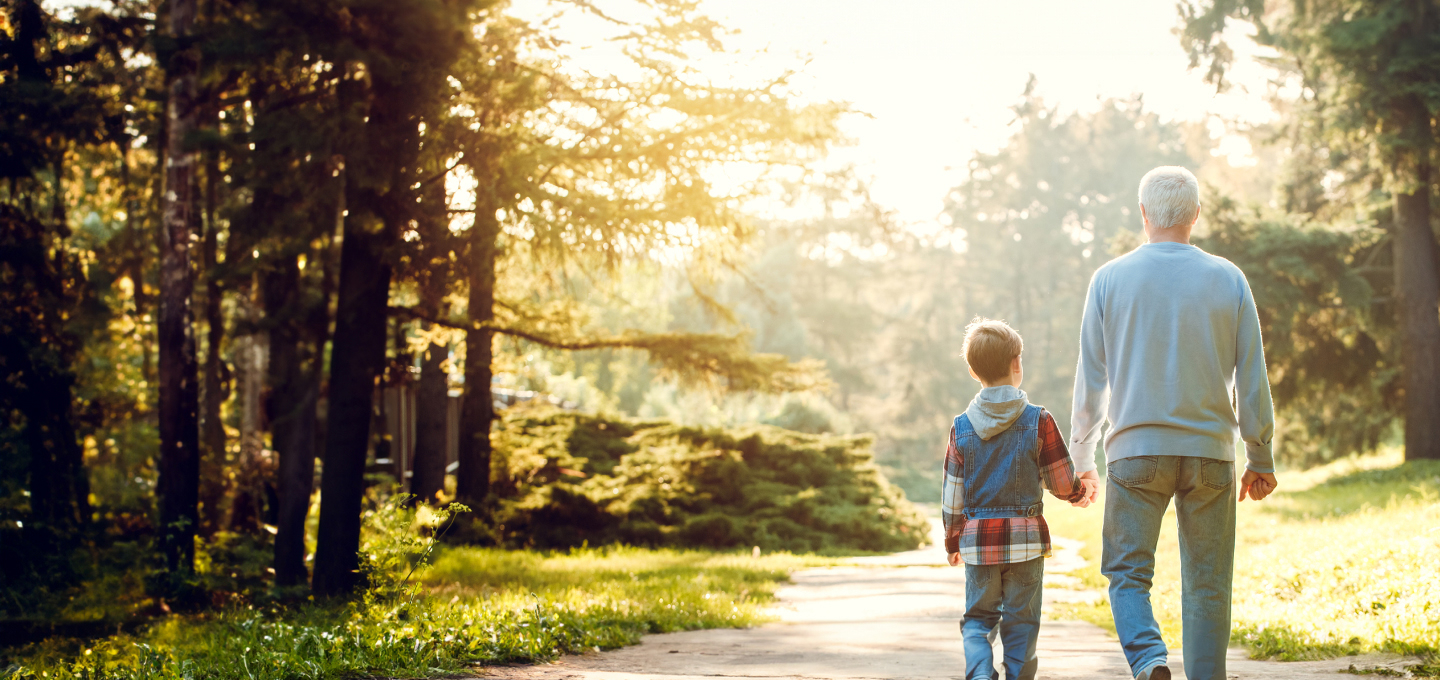 Imagem de pai e filho caminhando de mãos dadas e de costas. O pai é branco e usa um casaco azul e o filho é pequeno de pele branca, cabelos loiros e usa um casaco colorido. O pai está passando proteção e segurança ao filho.