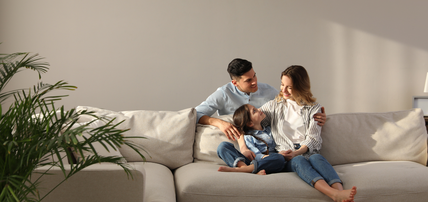 Imagem de uma família no sofá de casa. O pai é um homem branco e jovem, usando camisa azul. A mãe é uma mulher branca, jovem e de cabelos castanhos e camisa listrada. E a filha é uma criança branca e cabelos castanhos vestindo uma blusa azul. Eles estão sorridentes e se sentindo seguros em casa.