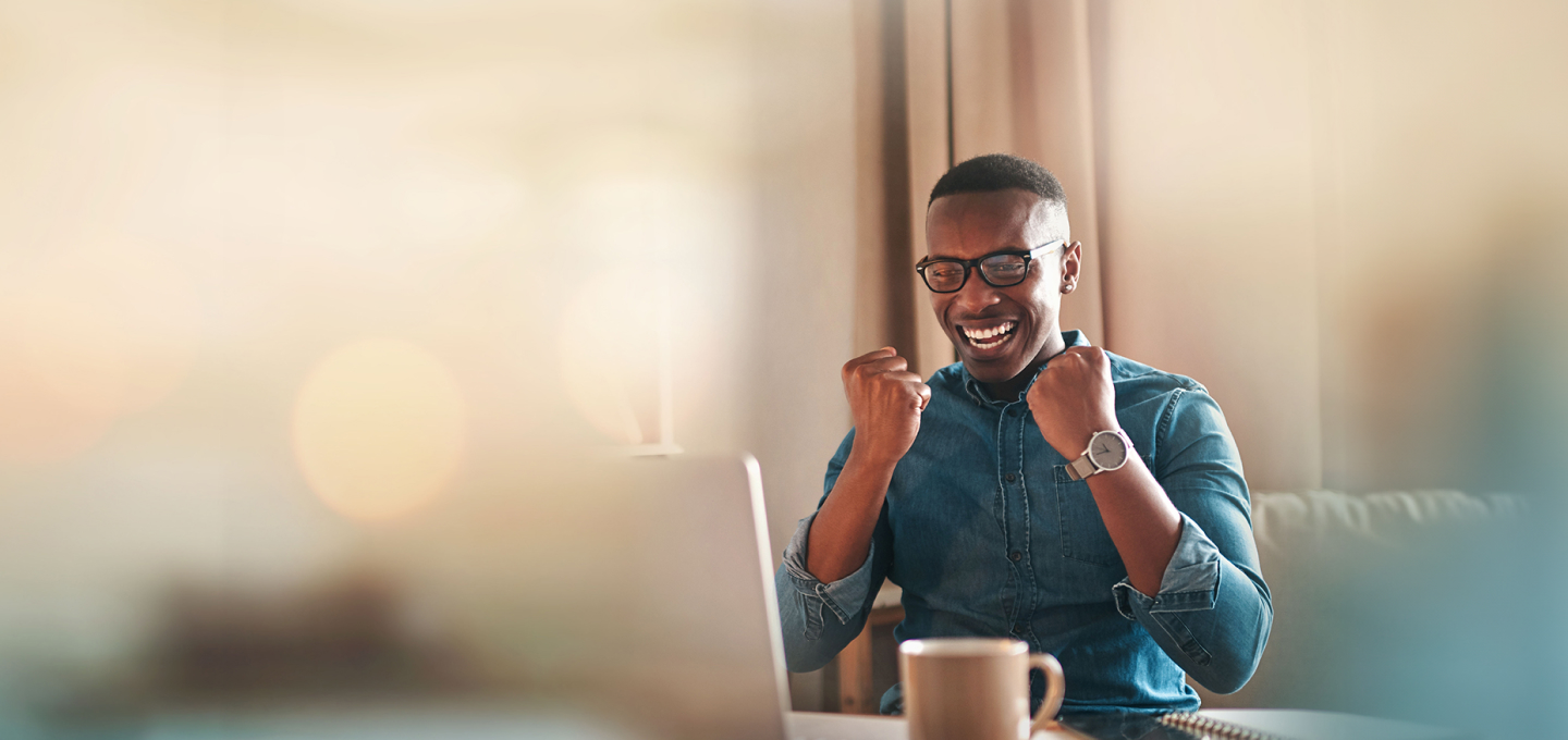 Imagem de um homem preto vestindo camisa azul que está em sua mesa conferindo uma mensagem no computador. Ele está feliz e comemorando por ter sido sorteado.