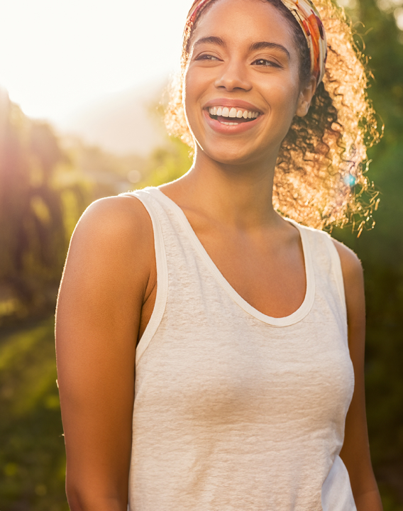 Imagem de uma mulher preta, com cabelos pretos cacheados em um local com árvores. Ela tem um sorriso bonito, está feliz e passa muita autoestima.