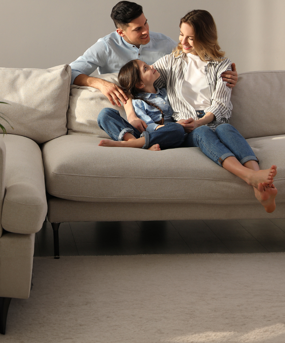 Imagem de uma família no sofá de casa. O pai é um homem branco e jovem, usando camisa azul. A mãe é uma mulher branca, jovem e de cabelos castanhos e camisa listrada. E a filha é uma criança branca e cabelos castanhos vestindo uma blusa azul. Eles estão sorridentes e se sentindo seguros em casa.