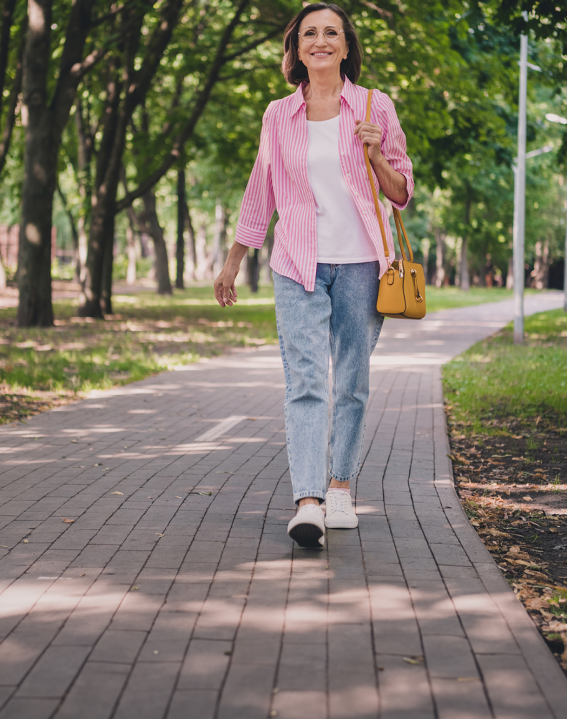 Imagem de uma mulher de meia idade andando em um parque durante o dia. Ela veste camisa rosa, jeans azul e carrega uma bolsa marrom com ar de proteção e tranquilidade.