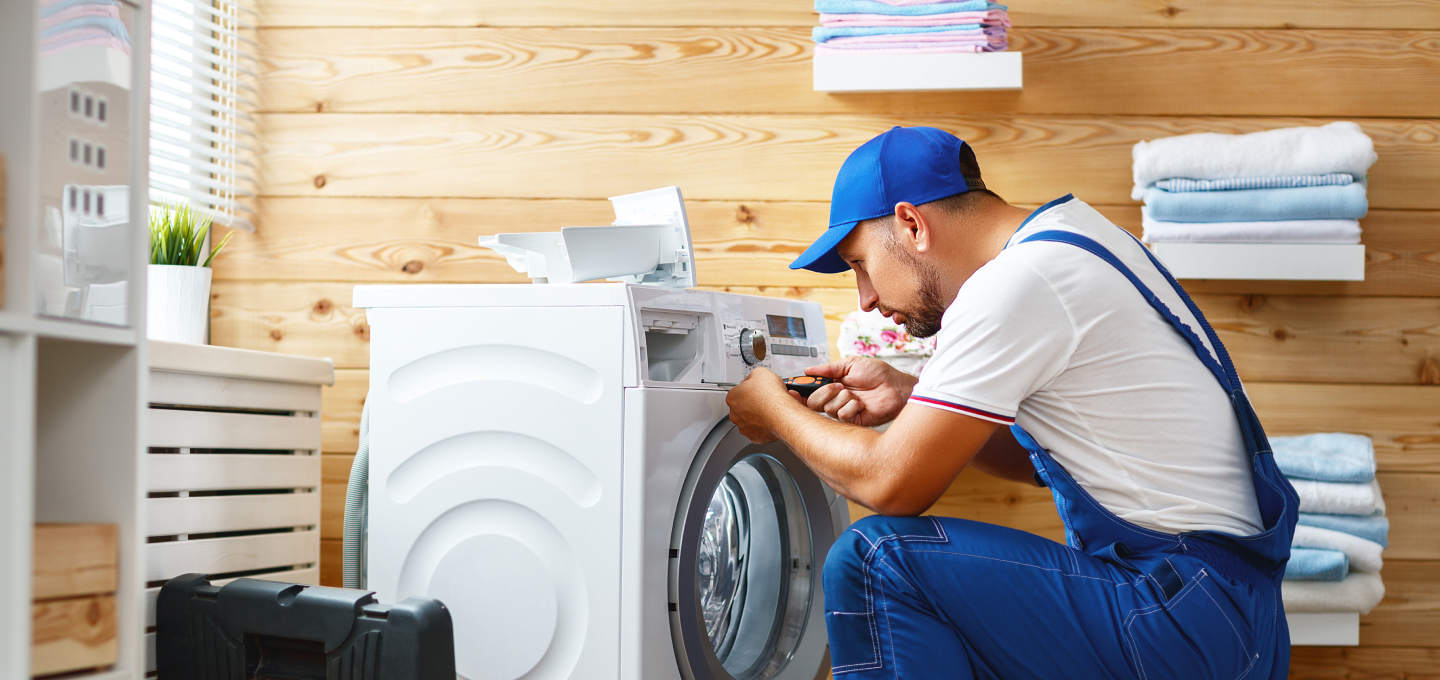 Imagem de um eletricista prestando assistência em uma casa e consertando uma máquina de lavar. Ele é branco com barba e veste uniforme e boné na cor azul.