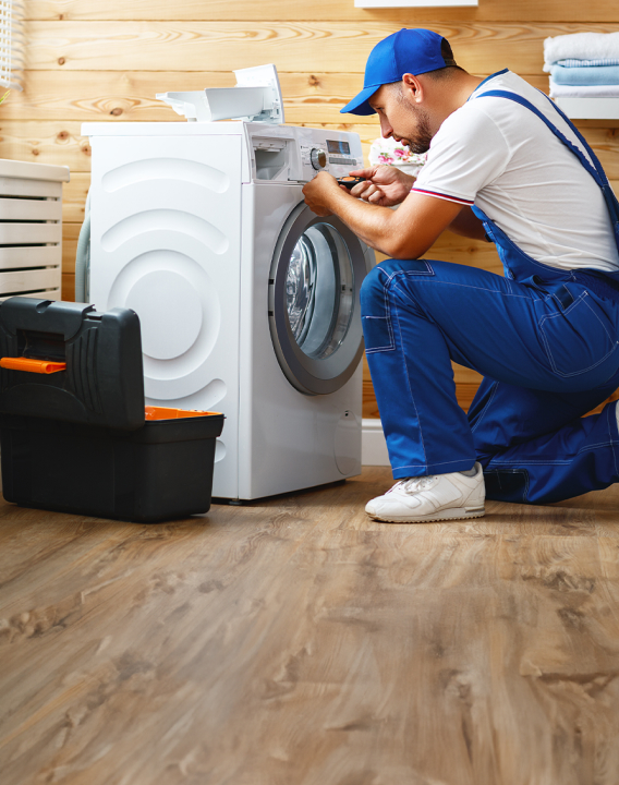 Imagem de um eletricista prestando assistência em uma casa e consertando uma máquina de lavar. Ele é branco com barba e veste uniforme e boné na cor azul.