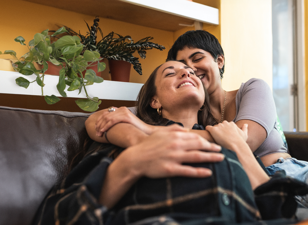 Duas mulheres jovens estão sentadas em um sofá marrom e se abraçam de forma carinhosa. Uma está com uma blusa na cor violeta e a outra com camisa xadrez preta. As duas usam piercing no nariz e sorriem muito à vontade. Atrás do sofá há uma estante com 3 vasos de plantas.