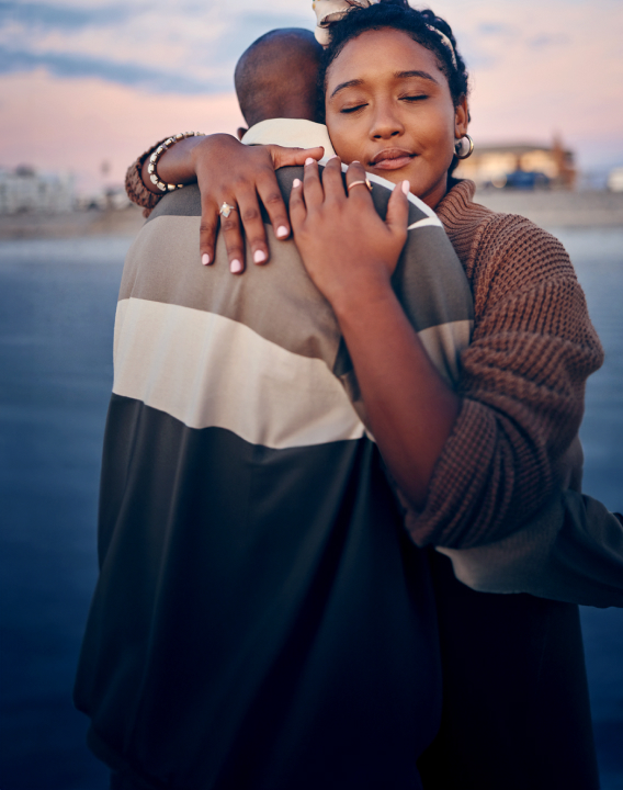 Imagem de um homem e de uma mulher se abraçando de forma carinhosa na beira de um lago. Ele é um homem preto, jovem e  usa camisa listrada. Ela, uma mulher preta, jovem e usa um casaco marrom. Eles se confortam e se amparam em um momento difícil.