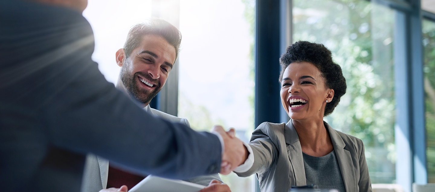 Pessoa sorridente apertando a mão de outra durante uma reunião profissional, enquanto um terceiro participante observa ao lado, em um ambiente corporativo iluminado por luz natural.